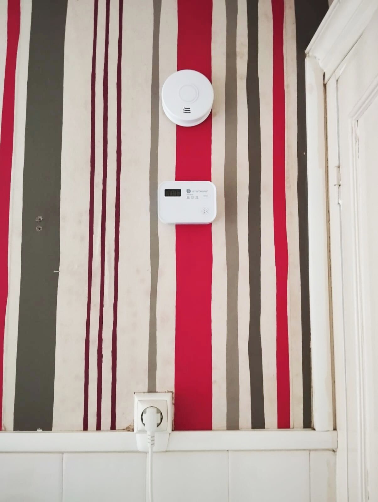 A wall with vertical stripes in red, white, and gray, a white device with a digital display, and a power outlet below.