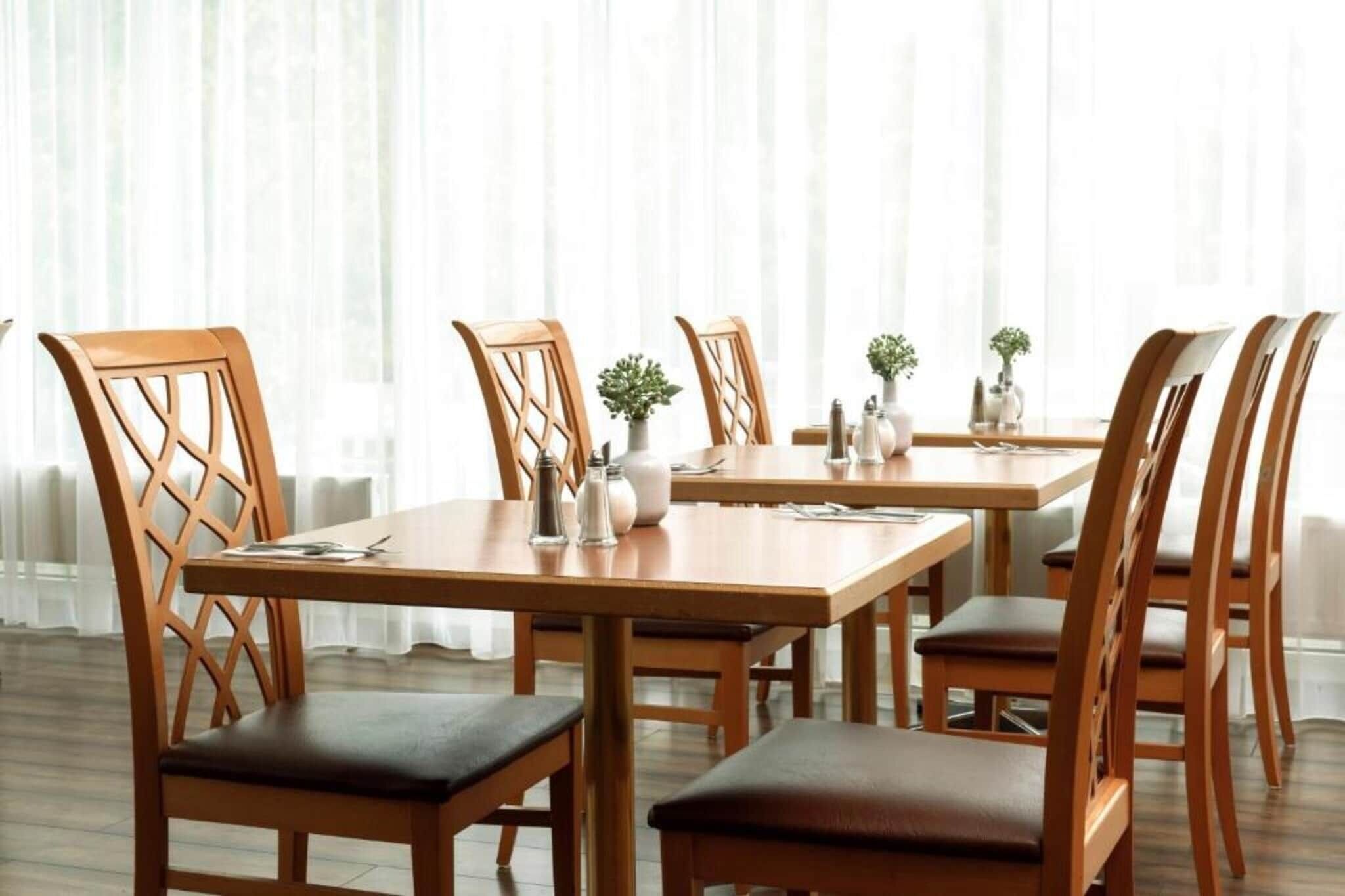 A dining area with wooden tables and chairs, a white curtain backdrop, and small potted plants on the tables.
