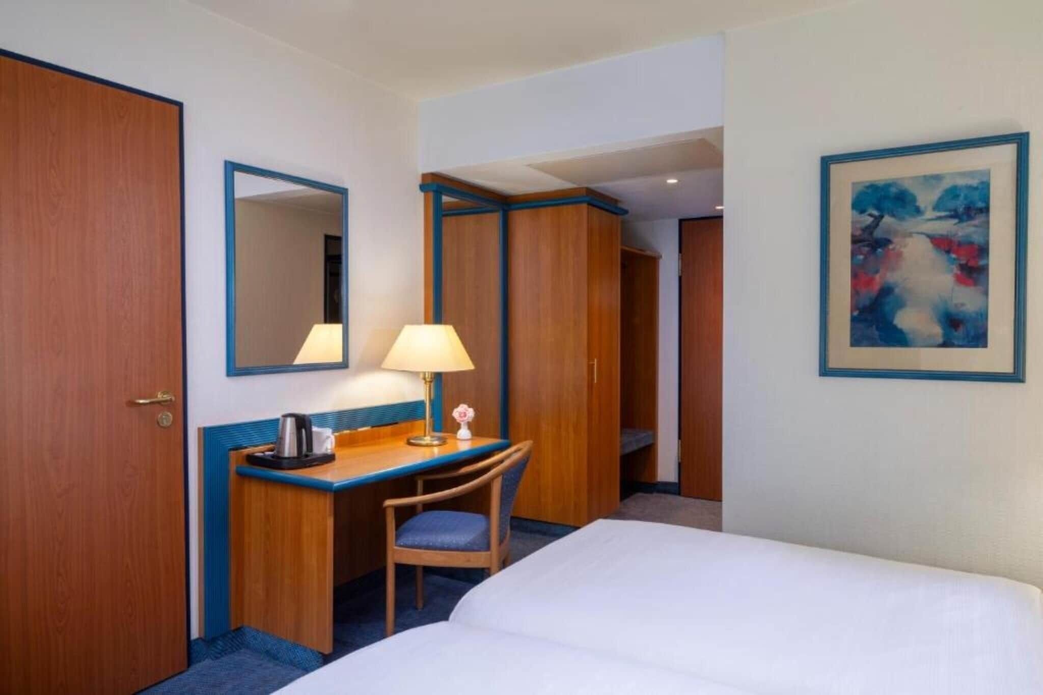 Een hotelkamer met een bed, een bureau met lamp, een stoel, een koffiezetapparaat, een spiegel en een schilderij aan de muur.