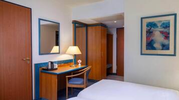 Een hotelkamer met een bed, een bureau met lamp, een stoel, een koffiezetapparaat, een spiegel en een schilderij aan de muur.