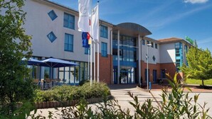 A hotel with a modern architectural design, featuring a central entrance with glass doors, a flagpole with multiple flags, and outdoor seating area with umbrellas. - Sunday Hotel Spa Wismar Disability access (Wismar)