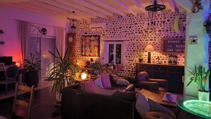 A cozy living room with a stone wall, a fireplace, and a dining area with a table and chairs. - Charming Air-Conditioned House –Pyrenees Views, Tennis & Large Garden (Sleeps 8) (Uzan)