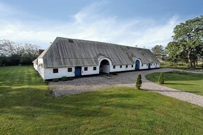 Un edificio bianco con tetto spiovente e ingresso ad arco, circondato da un prato curato e alberi.