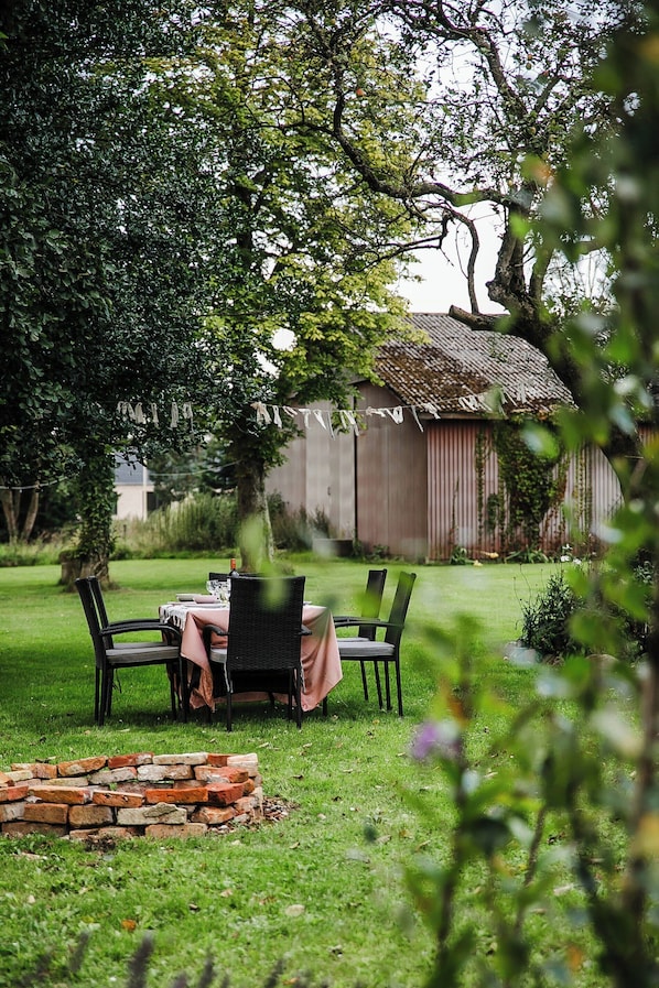 An outdoor dining area with a table and chairs, surrounded by greenery and a brick fire pit.