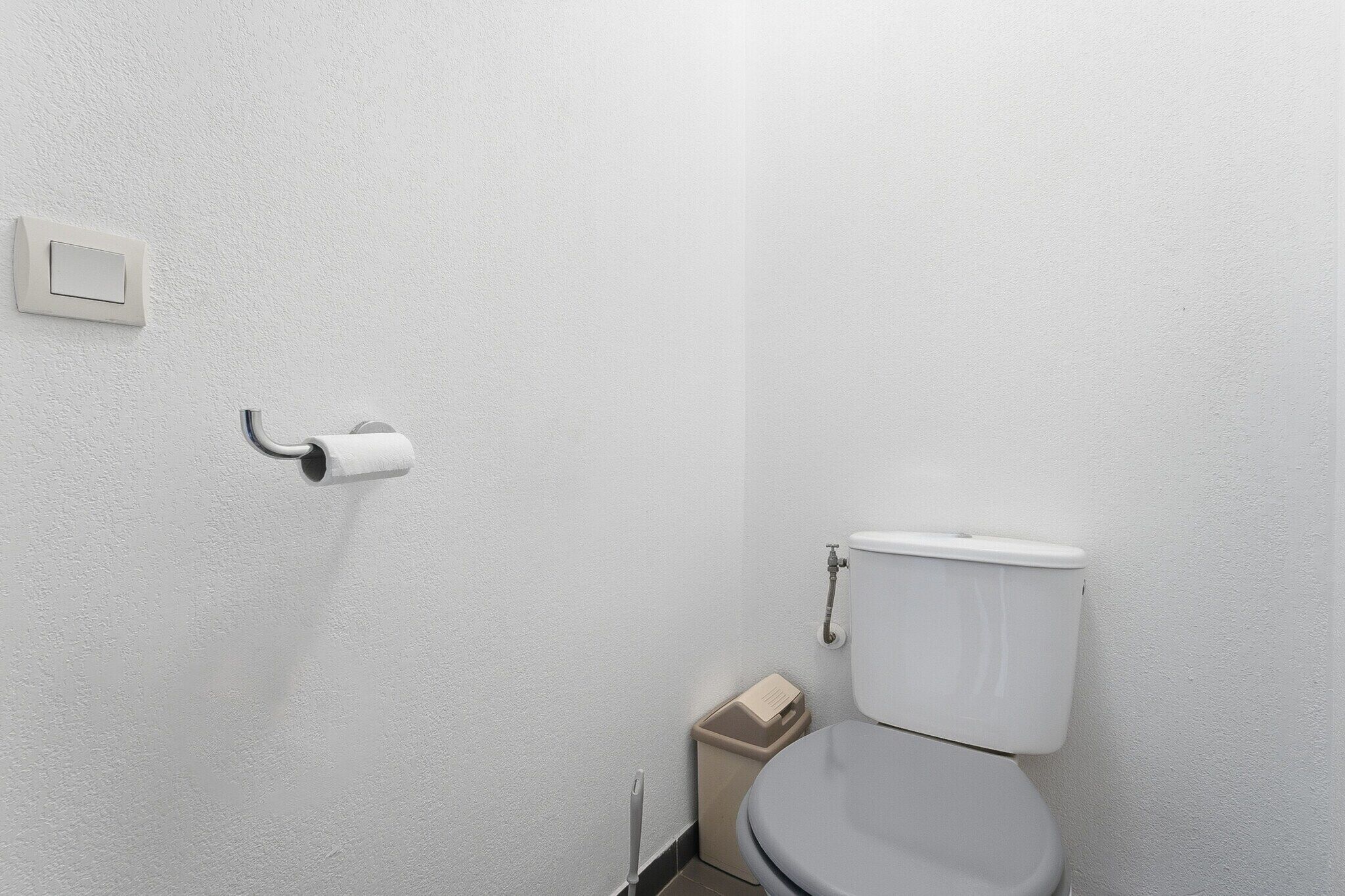 Eine weiße Toilette mit einem beigefarbenen Mülleimer daneben, einem Toilettenpapierhalter an der Wand und einem Spülhebel.