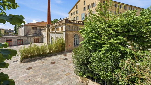Une cour intérieure avec un bâtiment doté de fenêtres en arc de cercle et d’une haute cheminée, le tout entouré de verdure.