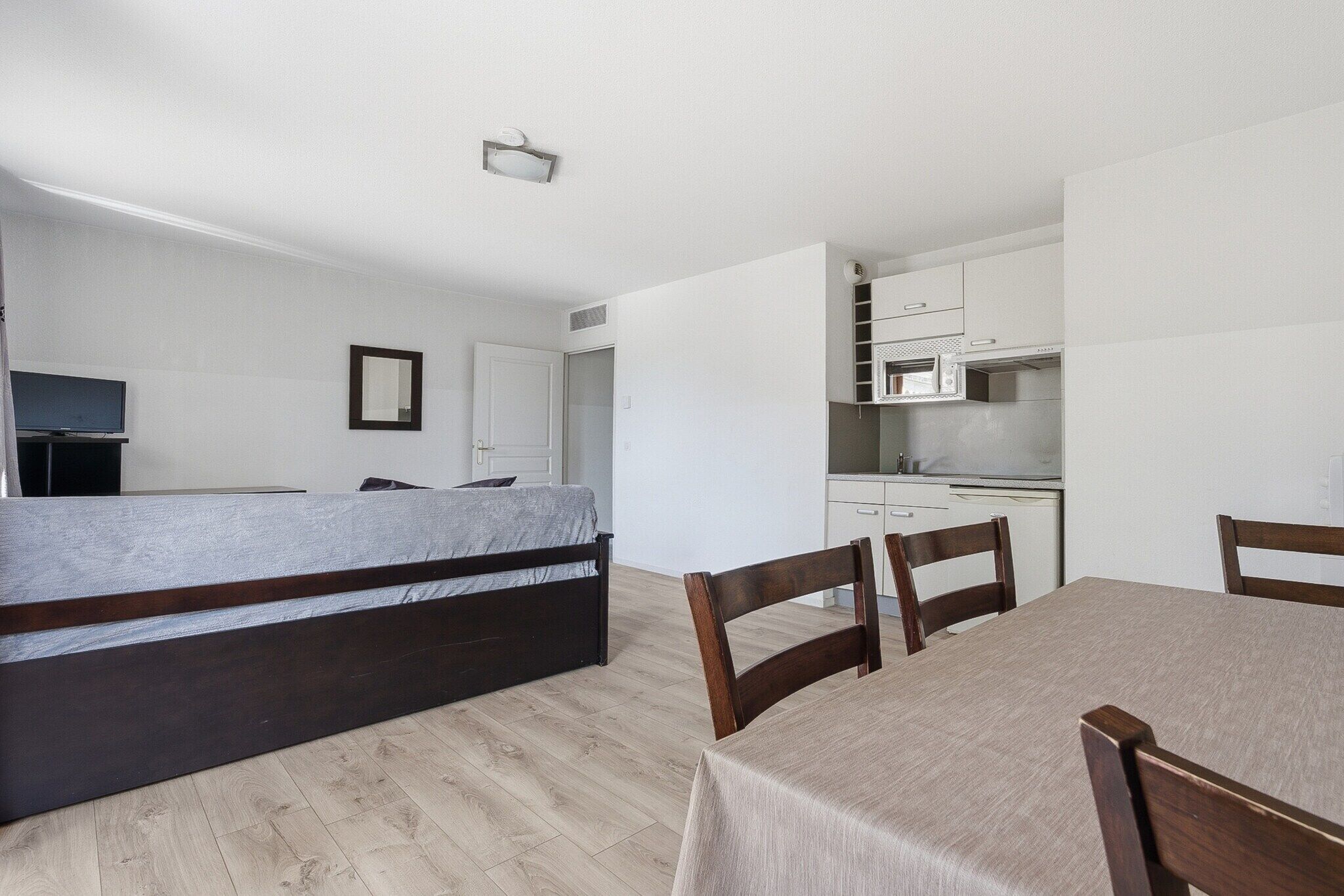 A bedroom with a bed, a dining table with chairs, a kitchenette, and a television.