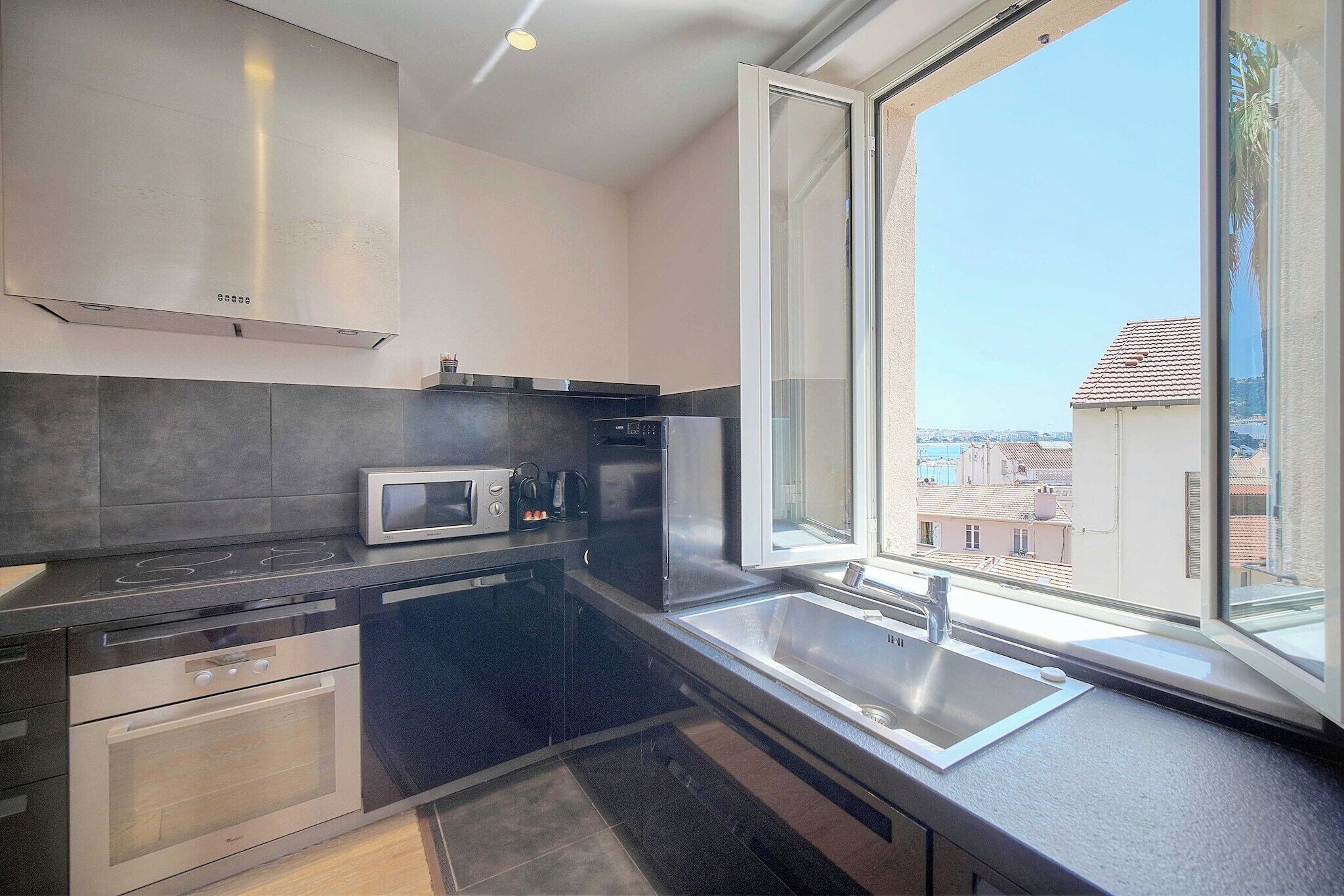 A modern kitchen with a built-in microwave, oven, and dishwasher, a large window with a view of buildings, and a sink.