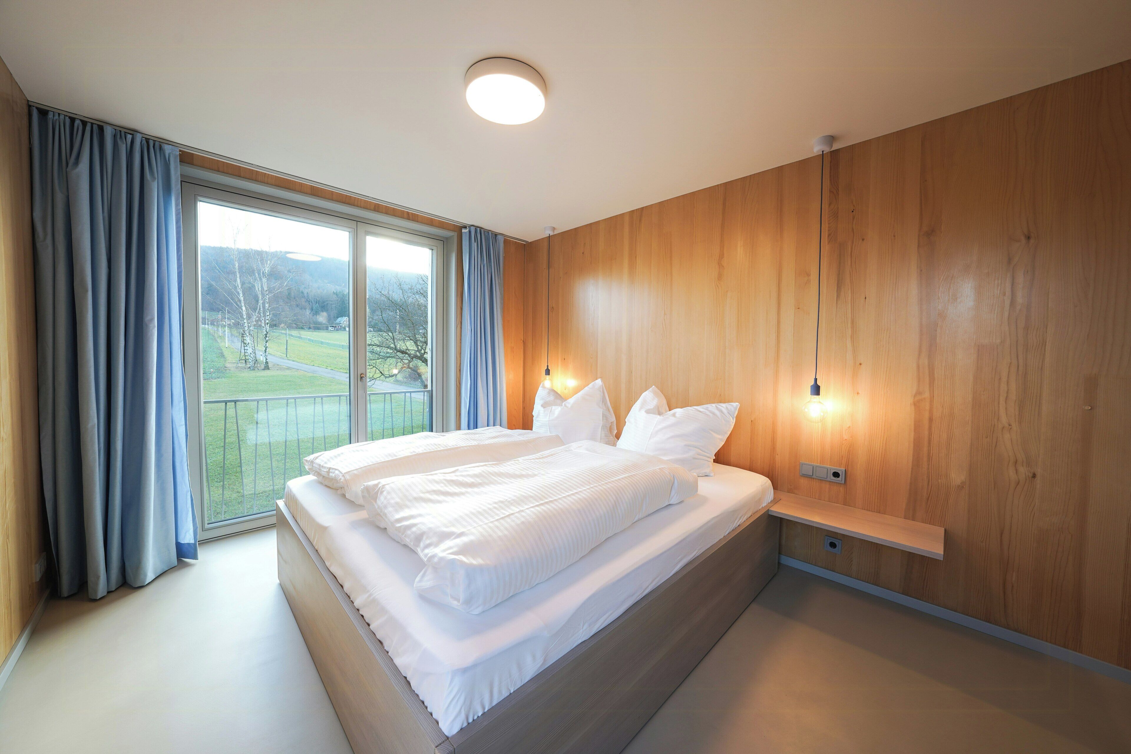 A modern bedroom with a large bed, a wooden wall, and a view of a green landscape through a large window.