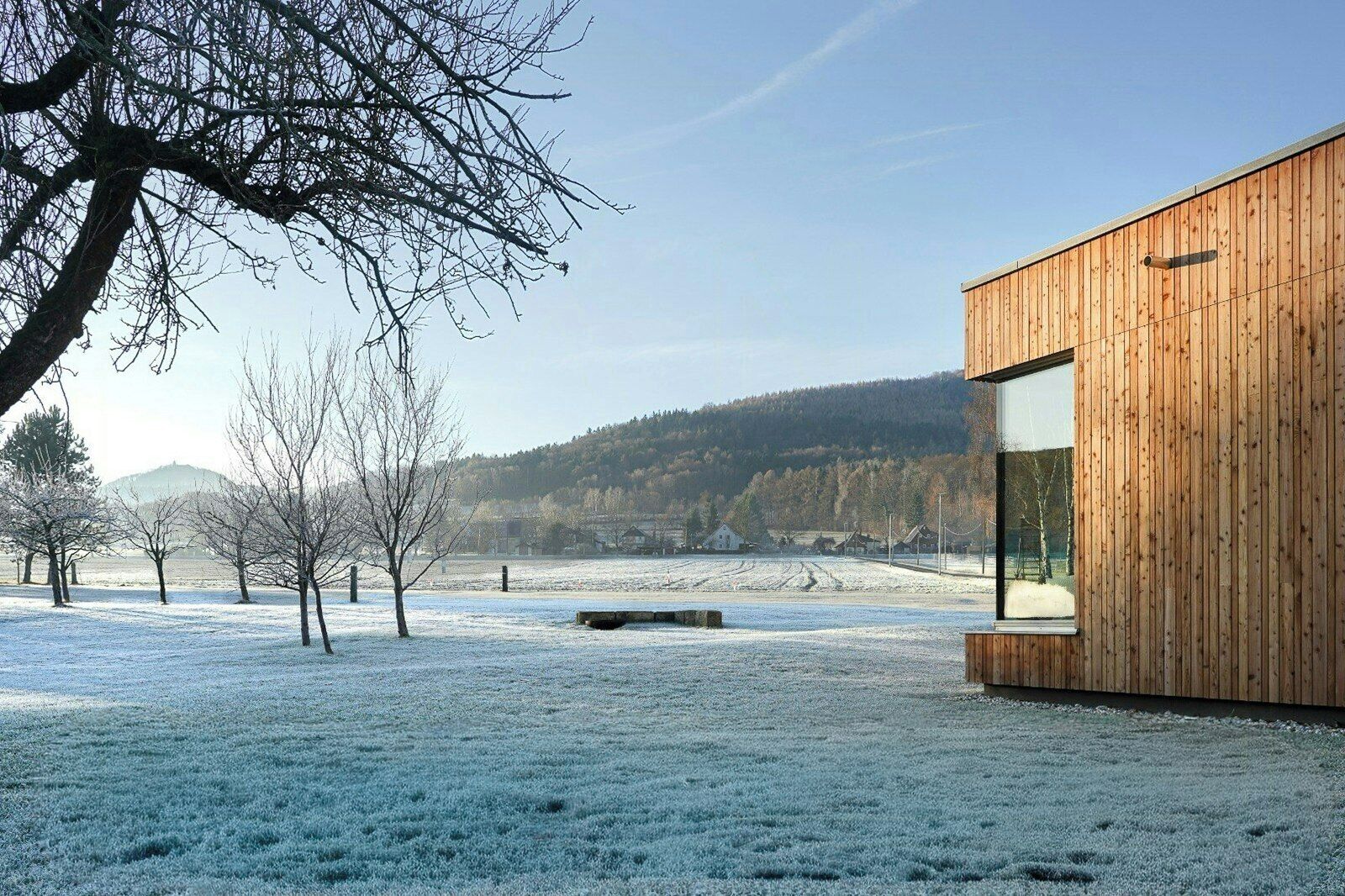 A modern building with a wooden facade and large windows, set in a snowy landscape with trees and a distant hill.