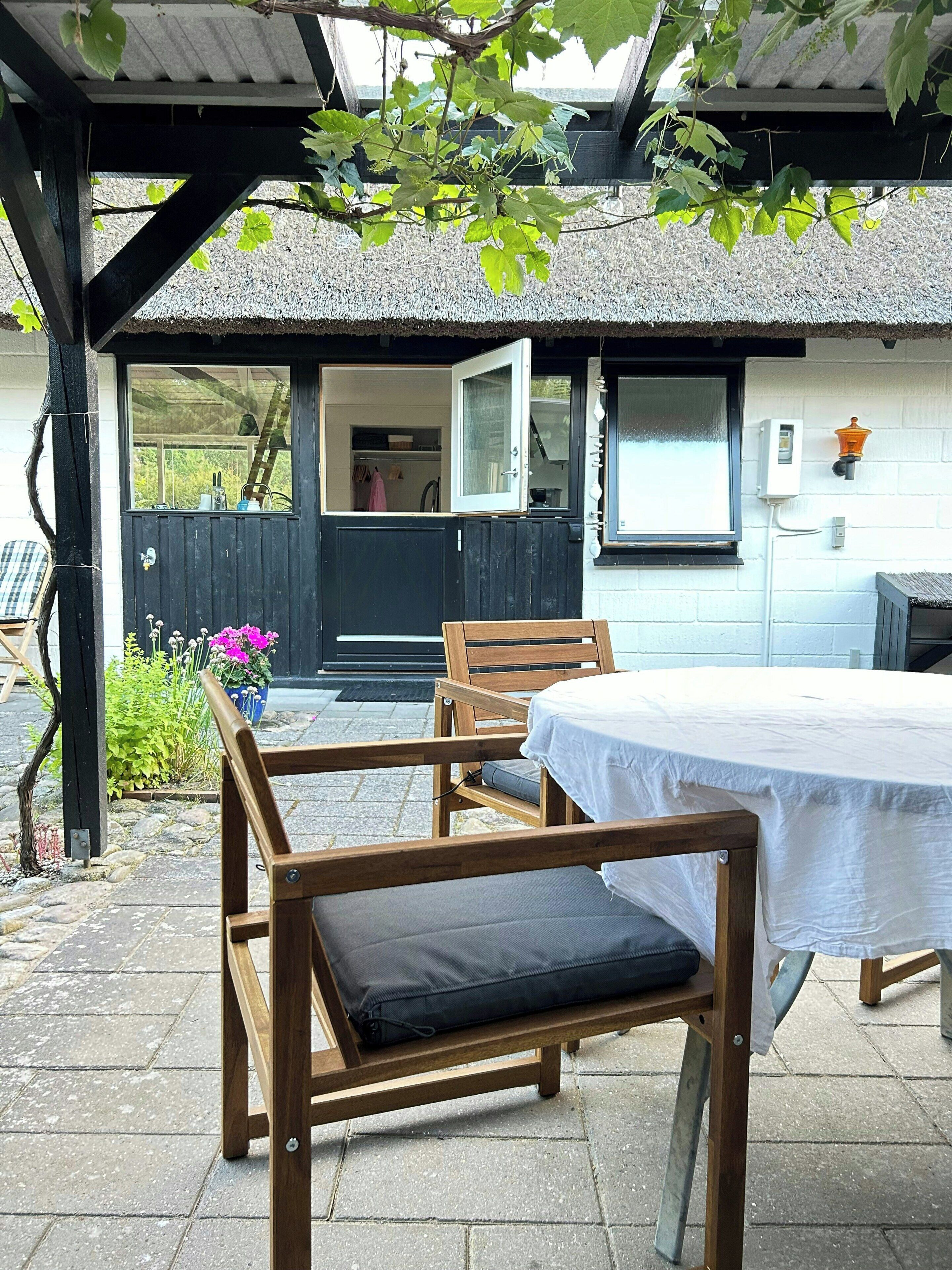 Een overdekt terras met een tafel en stoelen, een houten omheining en een deur die naar een kamer leidt.