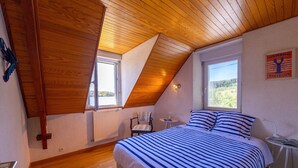 Ein Schlafzimmer mit schrägem Dach, einem Bett mit gestreifter Bettwäsche, einem Sessel, einem kleinen Tisch und einem Fenster mit Blick ins Grüne.