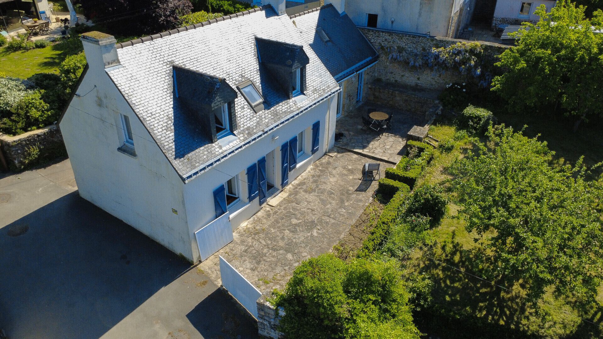 Un bâtiment blanc avec des volets bleus, un toit en pierre et une cour dallée.