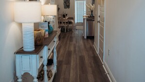 A hallway with a wooden floor, a white console table with a lamp, a mirror, and a door leading to a room. - Welcome to Easy Breezy.  Luxury Coastal Getaway, Minutes From Two Beaches. (Gulf Breeze)