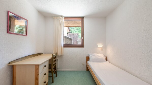 Una habitación pequeña y funcional con una cama individual, un escritorio con cajones, una silla, un ventanal con vistas a zonas verdes y una lámpara fijada a la pared.