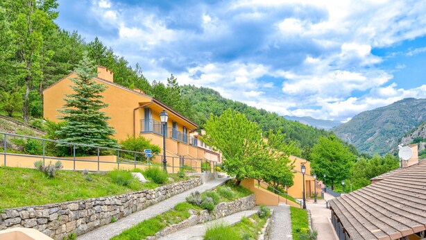 Una zona residencial en una ladera con escalones de piedra que conducen a un edificio con balcones, rodeado de vegetación y montañas al fondo.