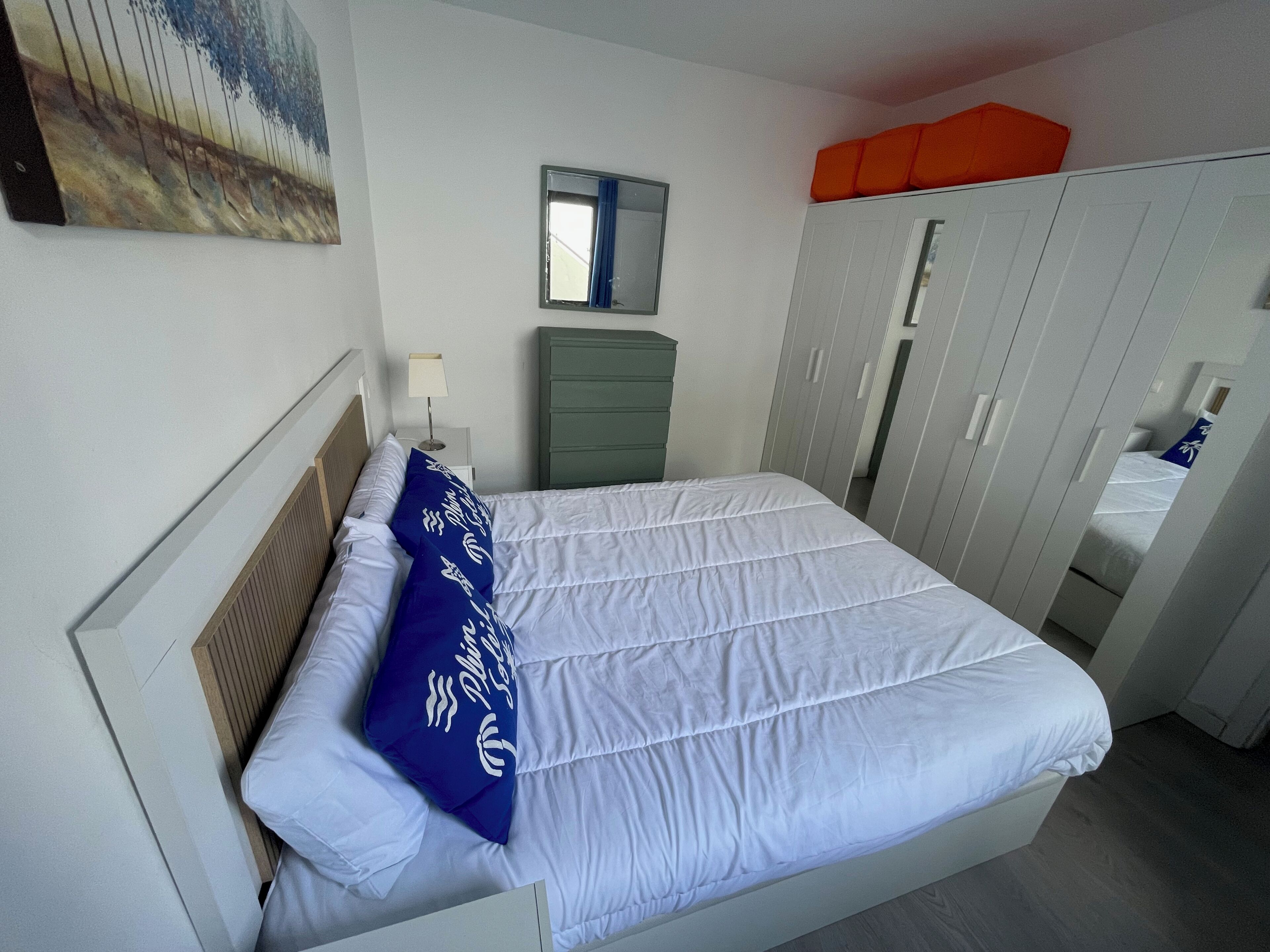A neatly made bed with white bedding and a blue pillow, a small bedside table with a lamp, a green dresser, and a wardrobe with a mirror.