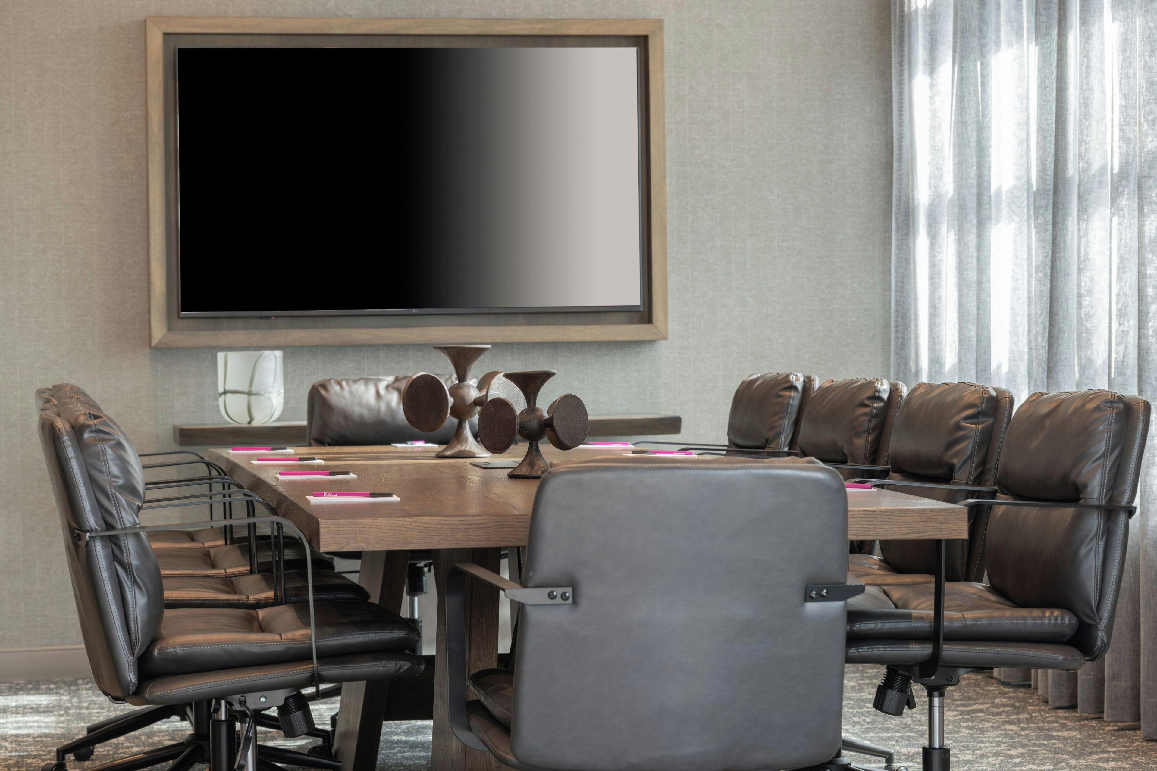 Une salle de conférence équipée d’un grand téléviseur à écran plat, de fauteuils en cuir, d’une table en bois et offrant une vue sur l’extérieur grâce à des rideaux.