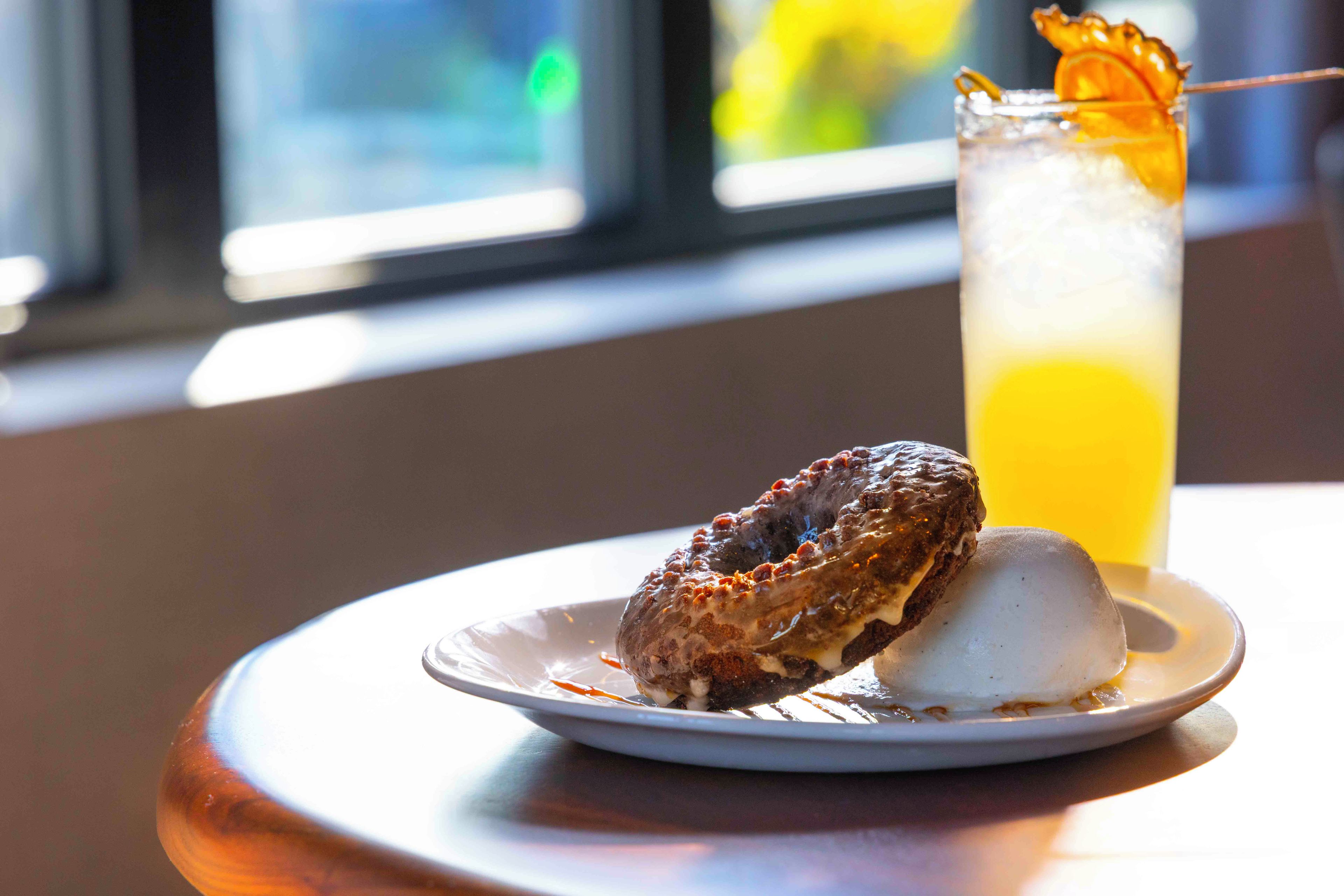 A dessert consisting of a donut and a scoop of ice cream on a plate, with a glass of iced beverage in the background.