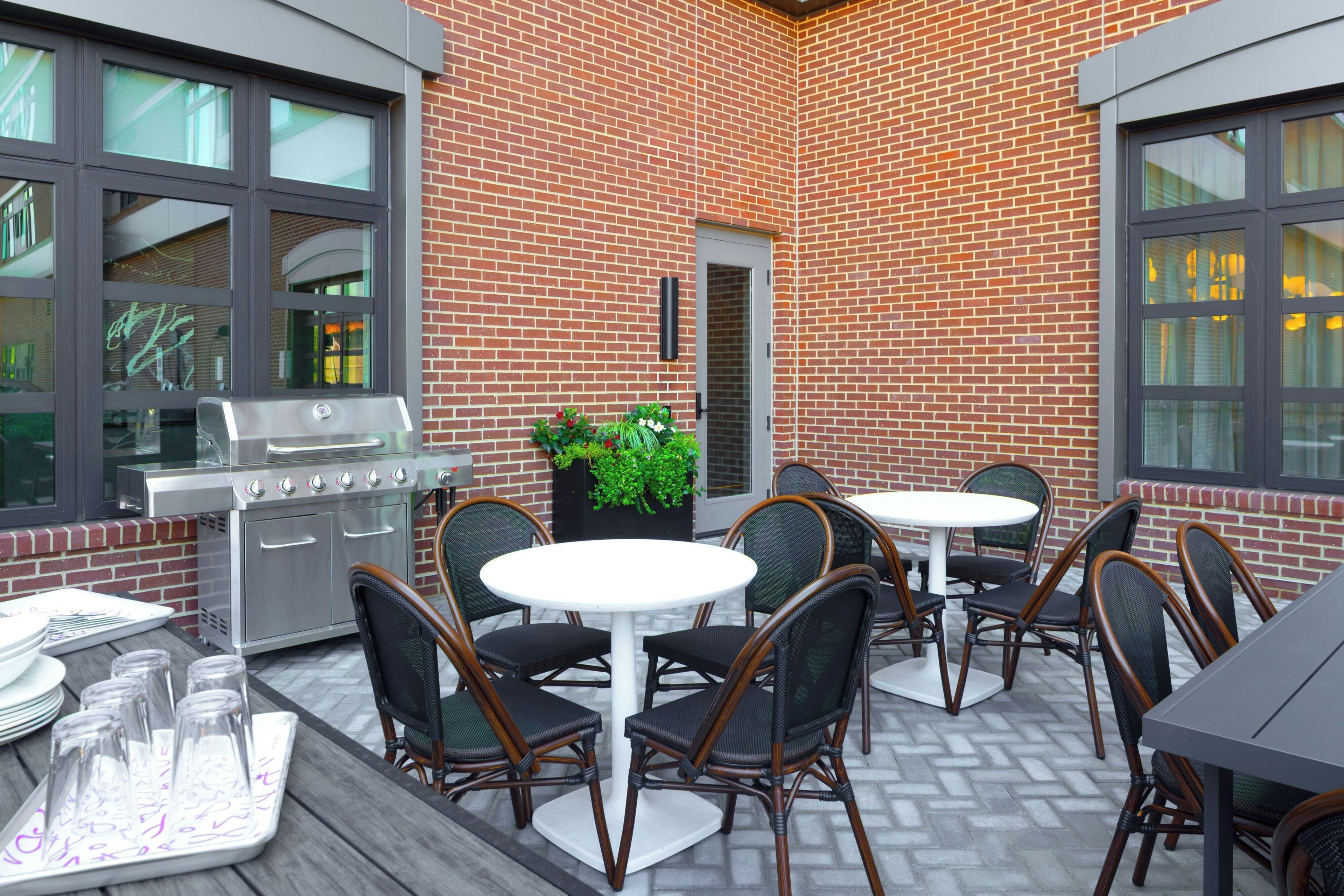 Un patio al aire libre con pared de ladrillo, una parrilla y mesas redondas con sillas.