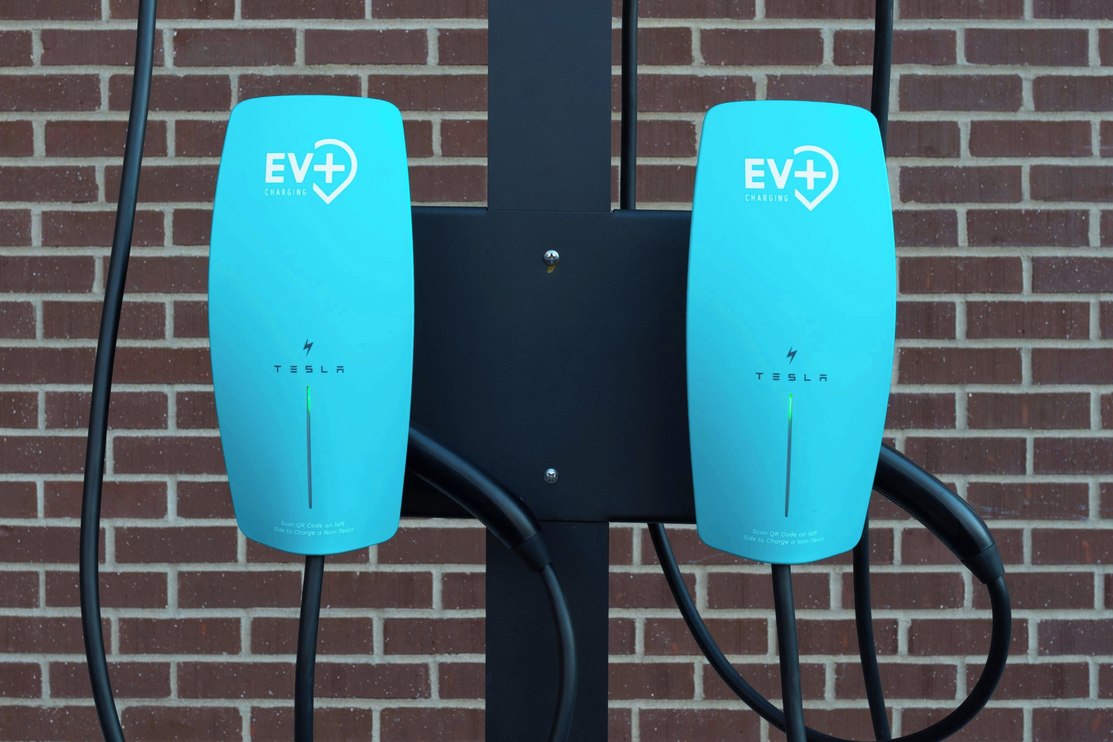 Dos estaciones de carga EV+ con la marca Tesla, conectadas a una pared de ladrillo.