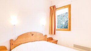 A bedroom with a wooden bed, bedside tables, a window with a view of a forest, and wall-mounted lights. - Alpine Apartment Near Slopes (Modane)