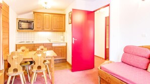 A compact kitchen with wooden cabinets, a microwave, and a dining area with a table and chairs. - Alpine Apartment Near Slopes (Modane)