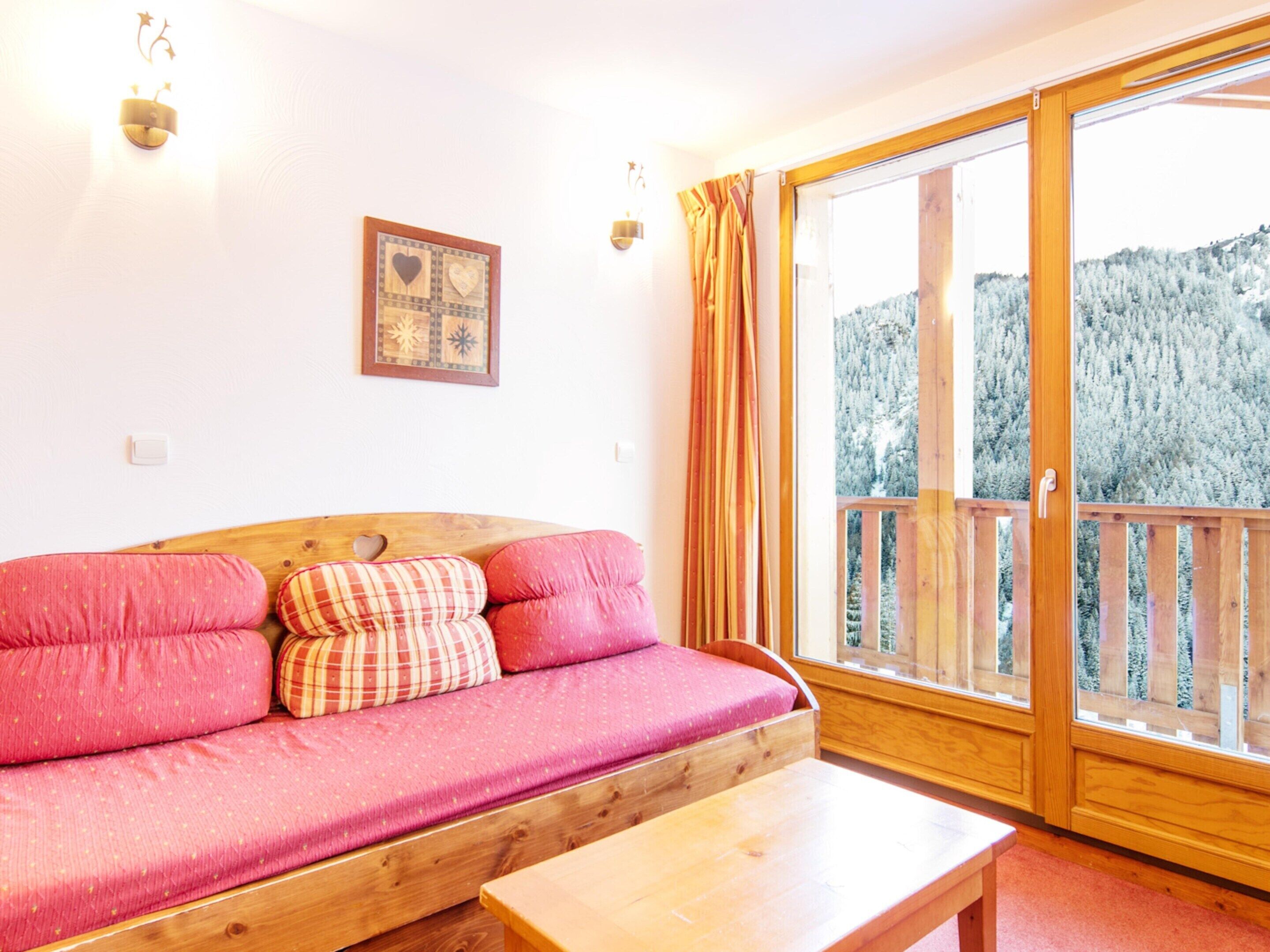 A wooden sofa with pink cushions, a wooden table, and a large window with a view of a forested mountain.