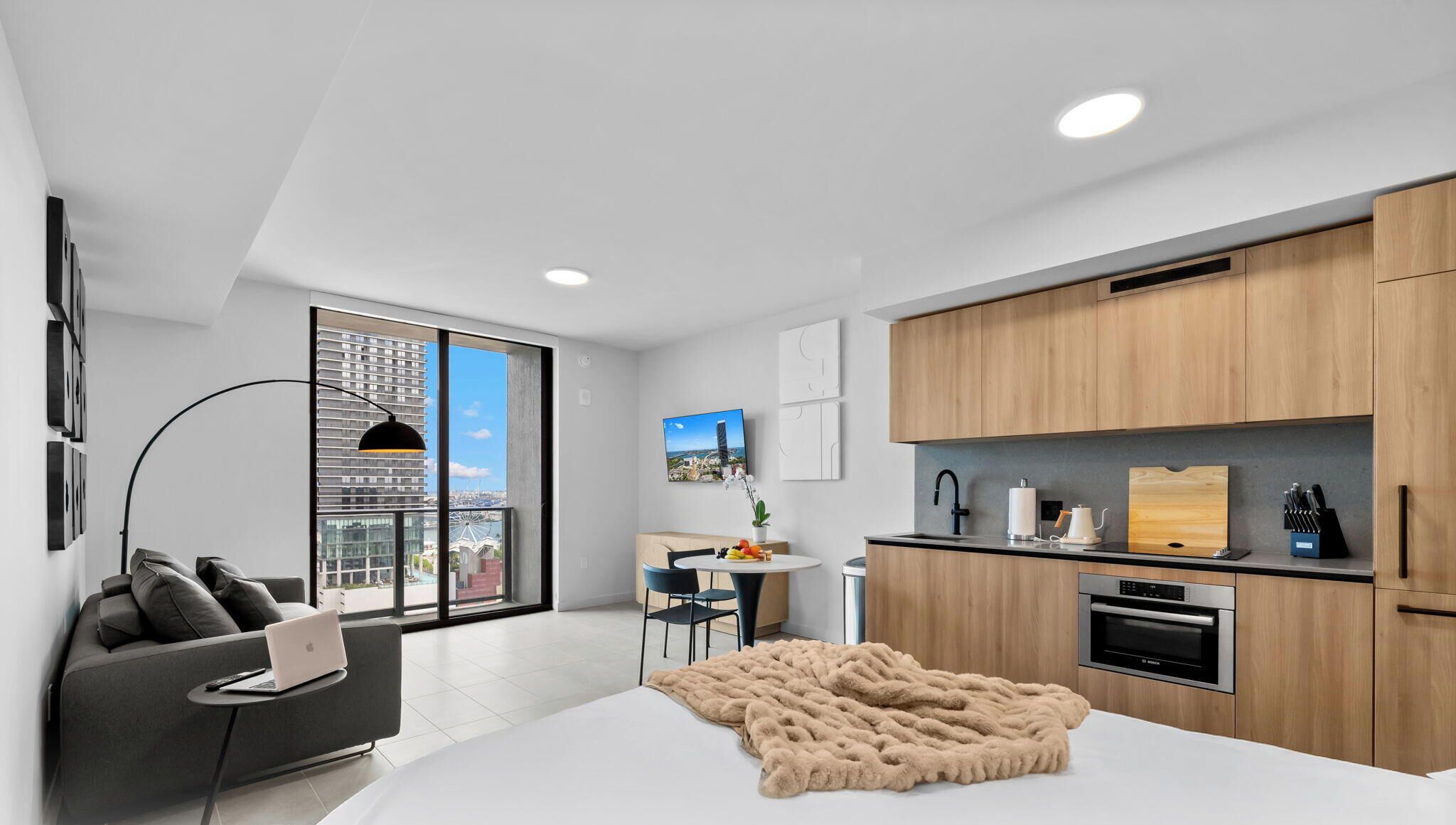A modern apartment with a large window, a dining area, a kitchenette, and a living space with a sofa and a laptop.
