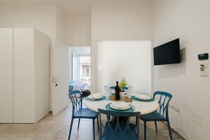 Een eetgedeelte met een ronde tafel, stoelen, een aan de muur bevestigde televisie en een deur naar een andere kamer.