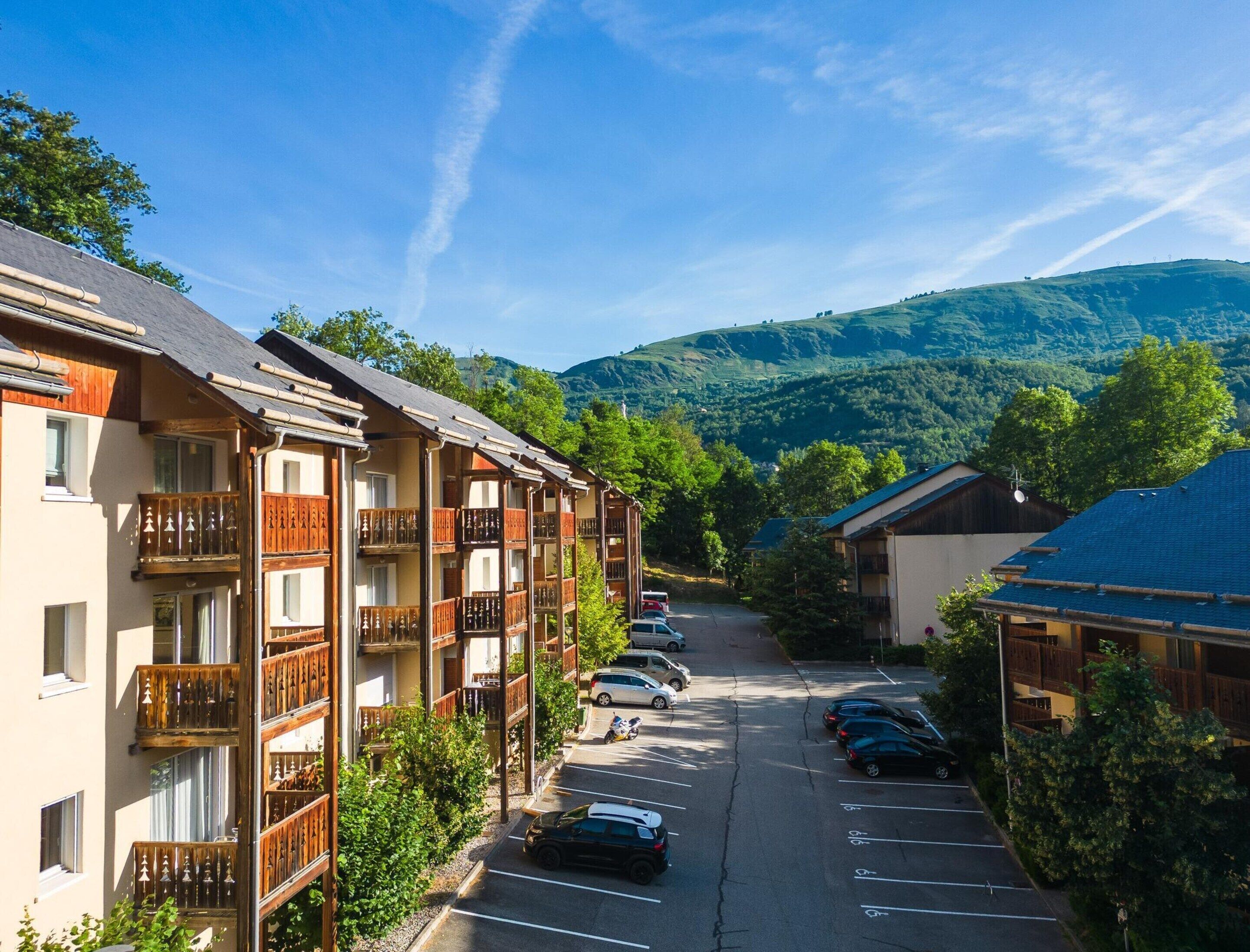 A row of multi-story buildings with balconies, a parking lot, and a mountainous backdrop.