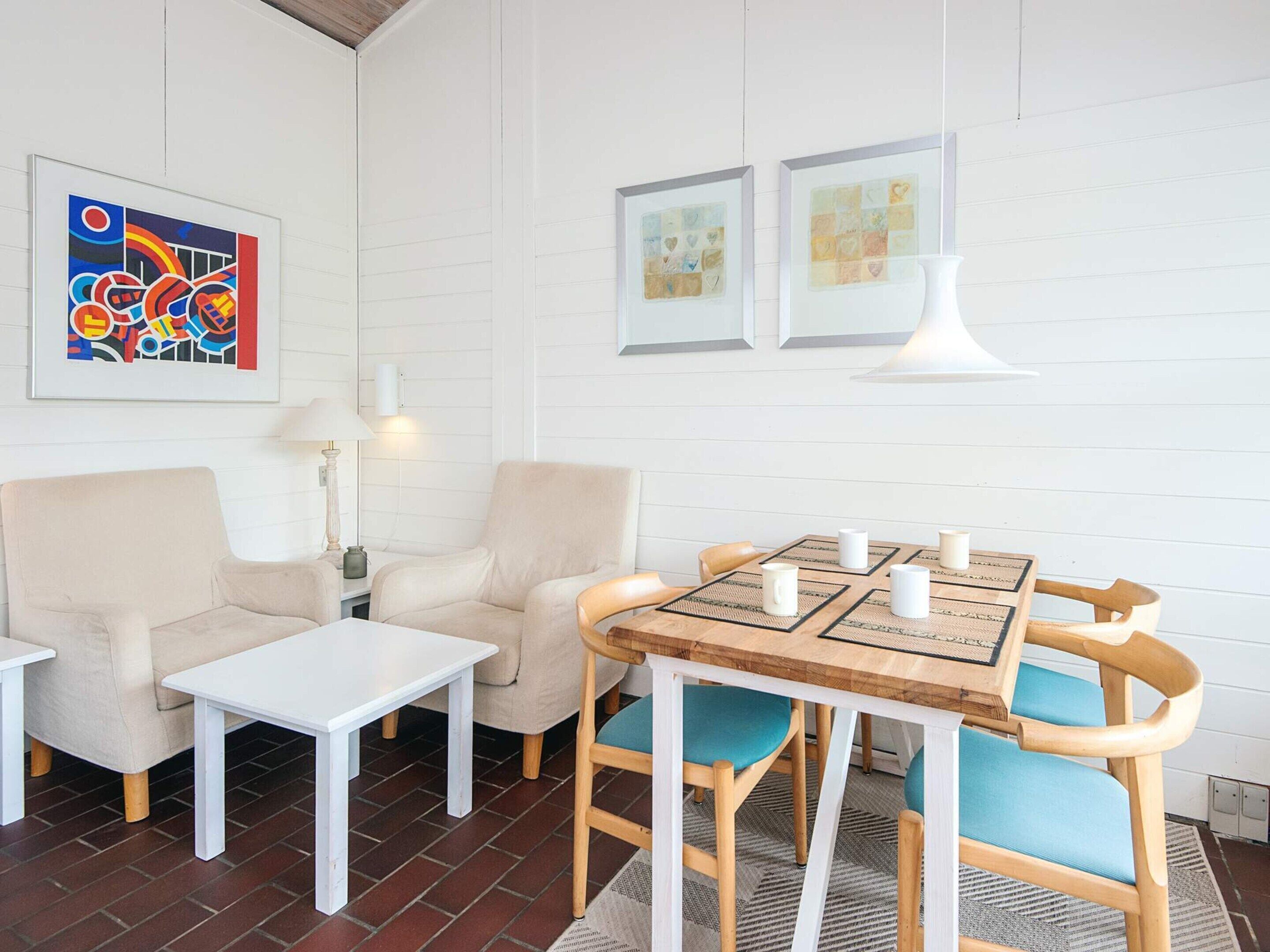 Een kamer met een witte baksteenmuur, een witte tafel met vier stoelen, een beige bank en een lamp.