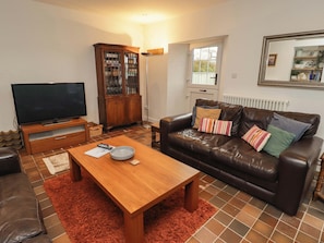 A living room with a brown leather sofa, a wooden coffee table, a television on a stand, a cabinet with glass doors, and a window with a view. - 8 Millmoor Farm Cottage (Haverfordwest)