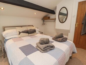 A neatly made bed with a quilt, pillows, and folded towels. There is a wooden shelf with a basket, a mirror on the wall, and a door with a towel hanging on it. - 8 Millmoor Farm Cottage (Haverfordwest)