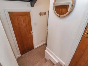 A narrow hallway with a wooden door, a carpeted floor, a white wall, and a round mirror. - 8 Millmoor Farm Cottage (Haverfordwest)