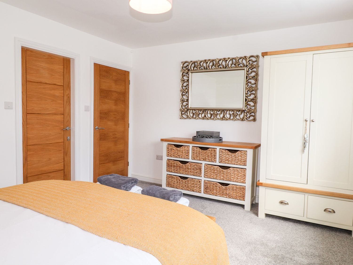 A bedroom with a bed, two wooden doors, a white wardrobe, a mirror, and wicker baskets on a white dresser.