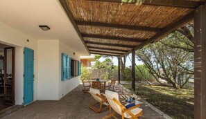 A covered outdoor area with a table and chairs, a door, and a view of trees and a building in the background. - Casa Sottotorre Beachfront (Calasetta)