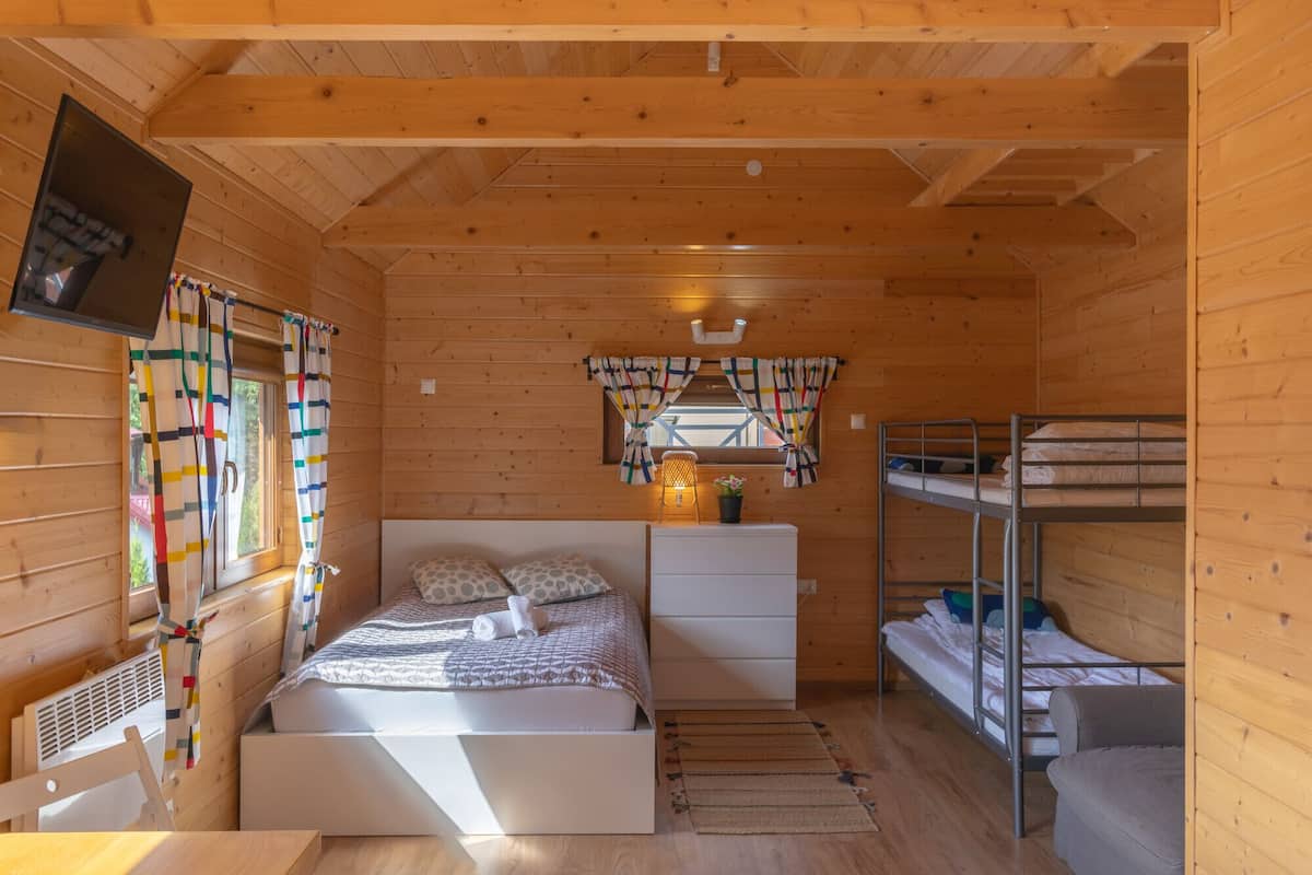A wooden cabin with a bunk bed, a single bed, a small table, a chair, a window with curtains, and a wall-mounted TV.