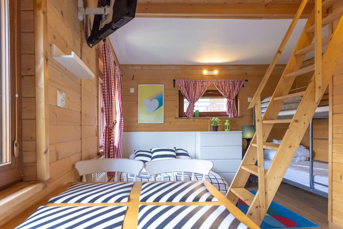 A wooden cabin interior with a bed, a desk, a ladder, and a window with curtains.