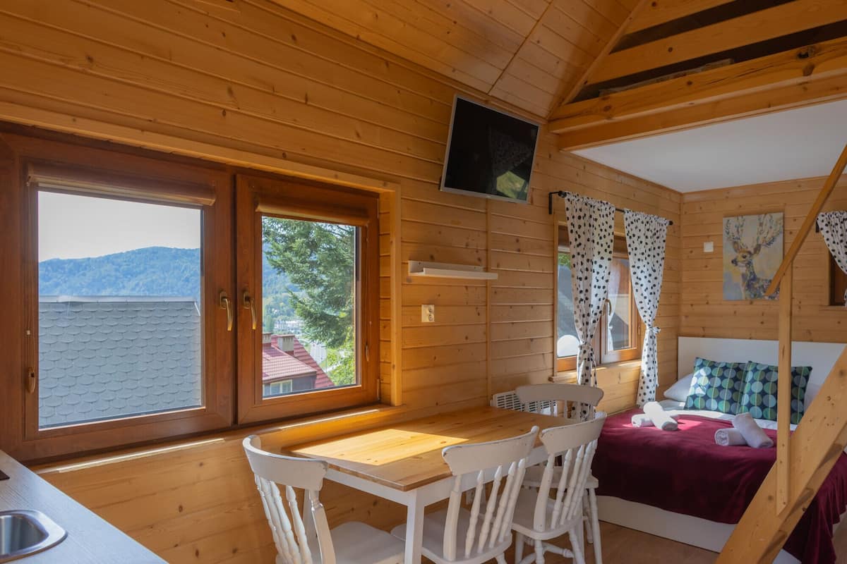 A cabin with a wooden interior, a dining area with a table and chairs, a bed with a red blanket, and a window with a view of a mountain.