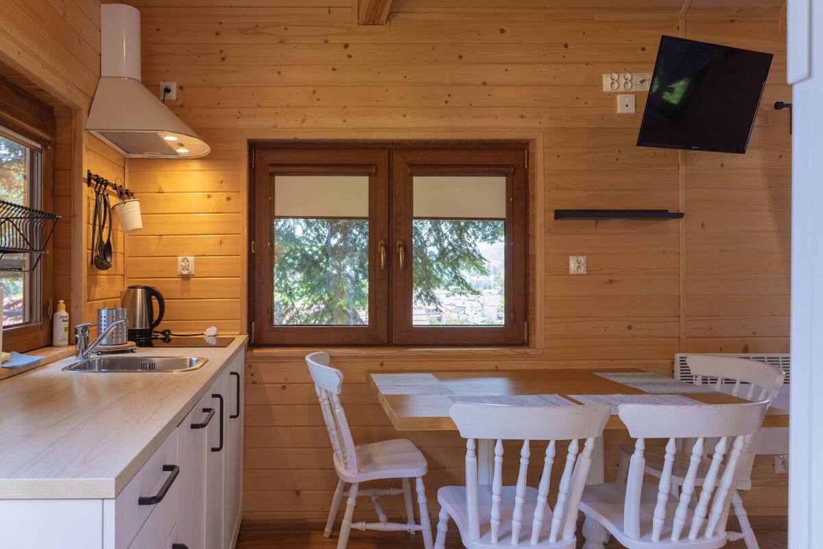 A wooden cabin interior with a kitchen area, two windows, a dining table with chairs, and a flat-screen TV mounted on the wall.