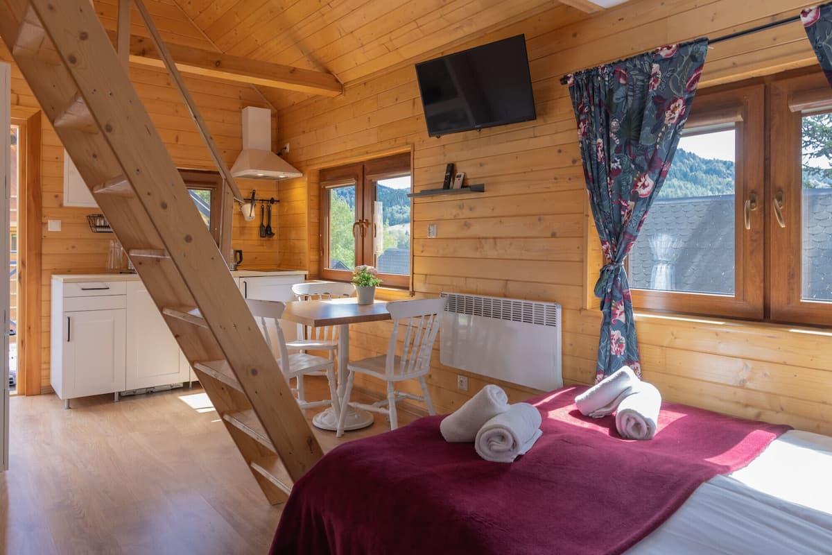 A wooden cabin interior with a bed, a dining table, a kitchen area, and a TV mounted on the wall.