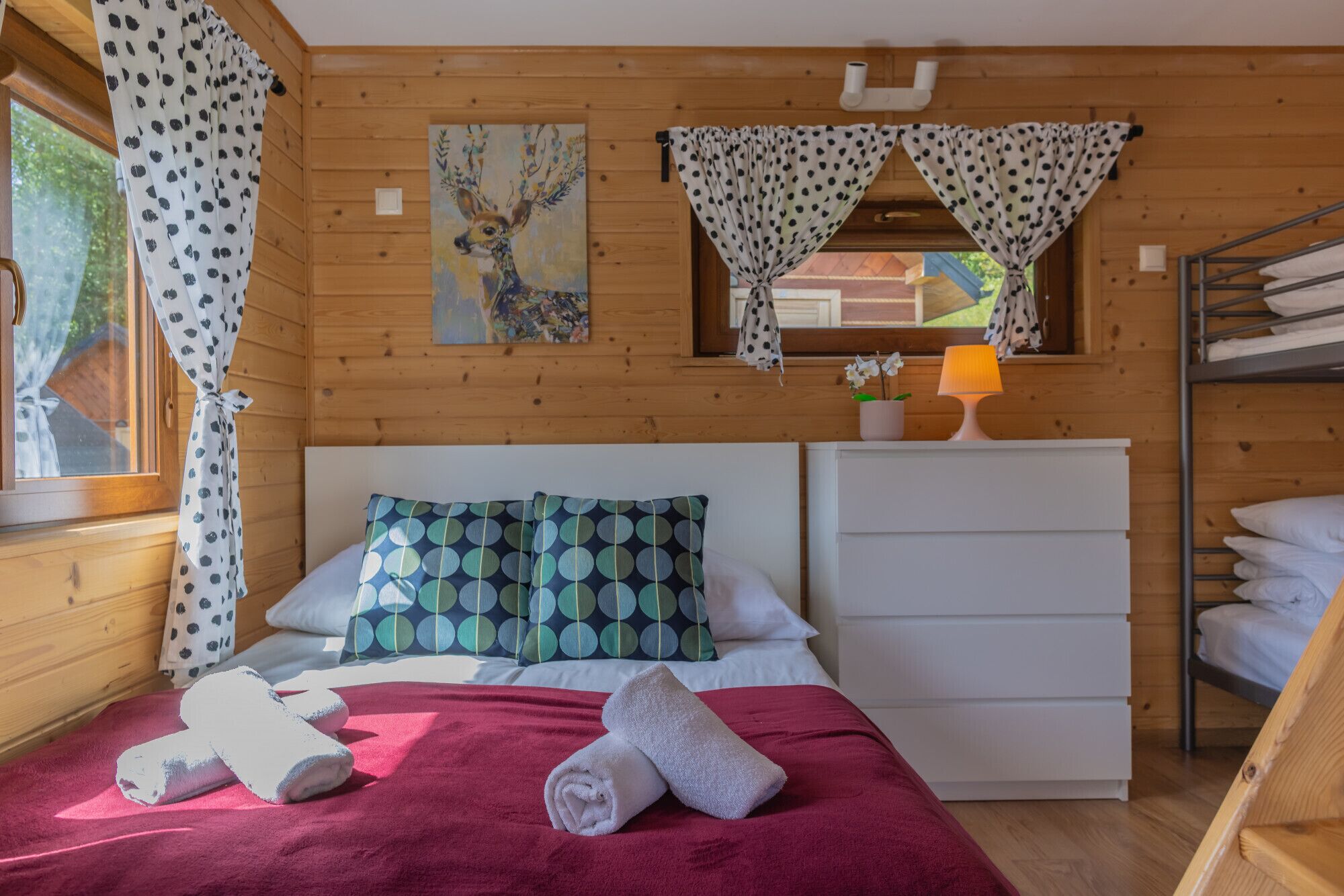 A wooden cabin bedroom with a bed, pillows, a red blanket, towels, a lamp, and a painting on the wall.
