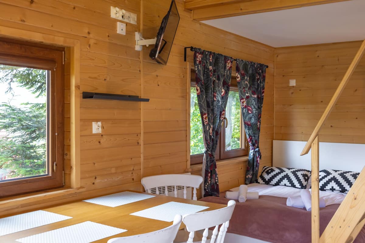 A wooden cabin room with a bed, a table, chairs, and a window with curtains.
