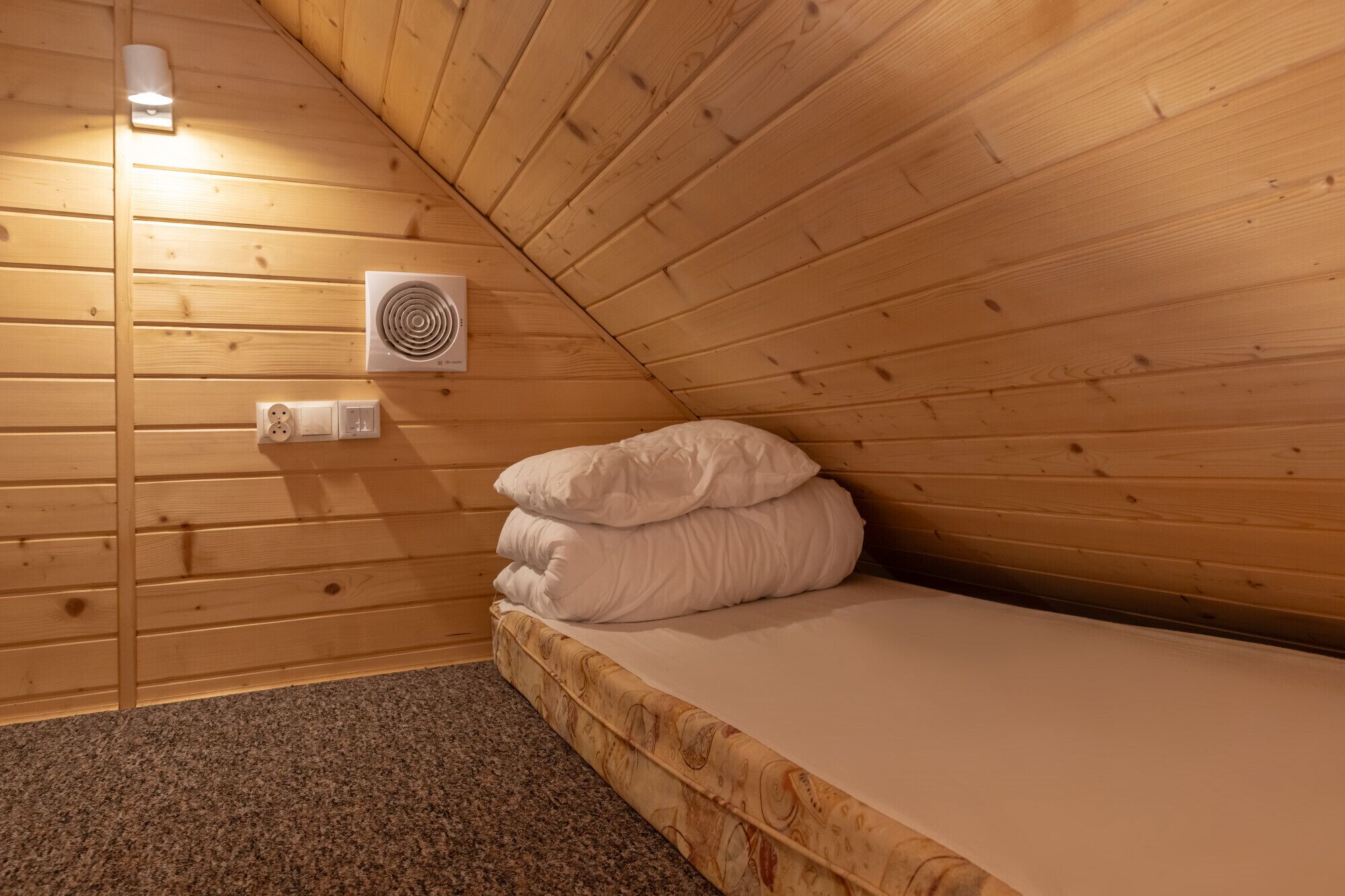Una habitación pequeña y acogedora con techo inclinado de madera, una cama individual con ropa blanca, un aire acondicionado de pared y un enchufe.