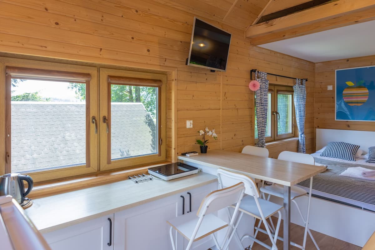 A wooden cabin interior with a kitchen area, dining table, chairs, and a bed.