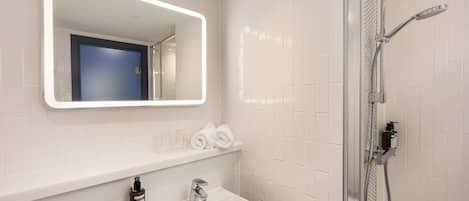 A bathroom with a white sink, a mirror with a light, a shower, and a blue door.