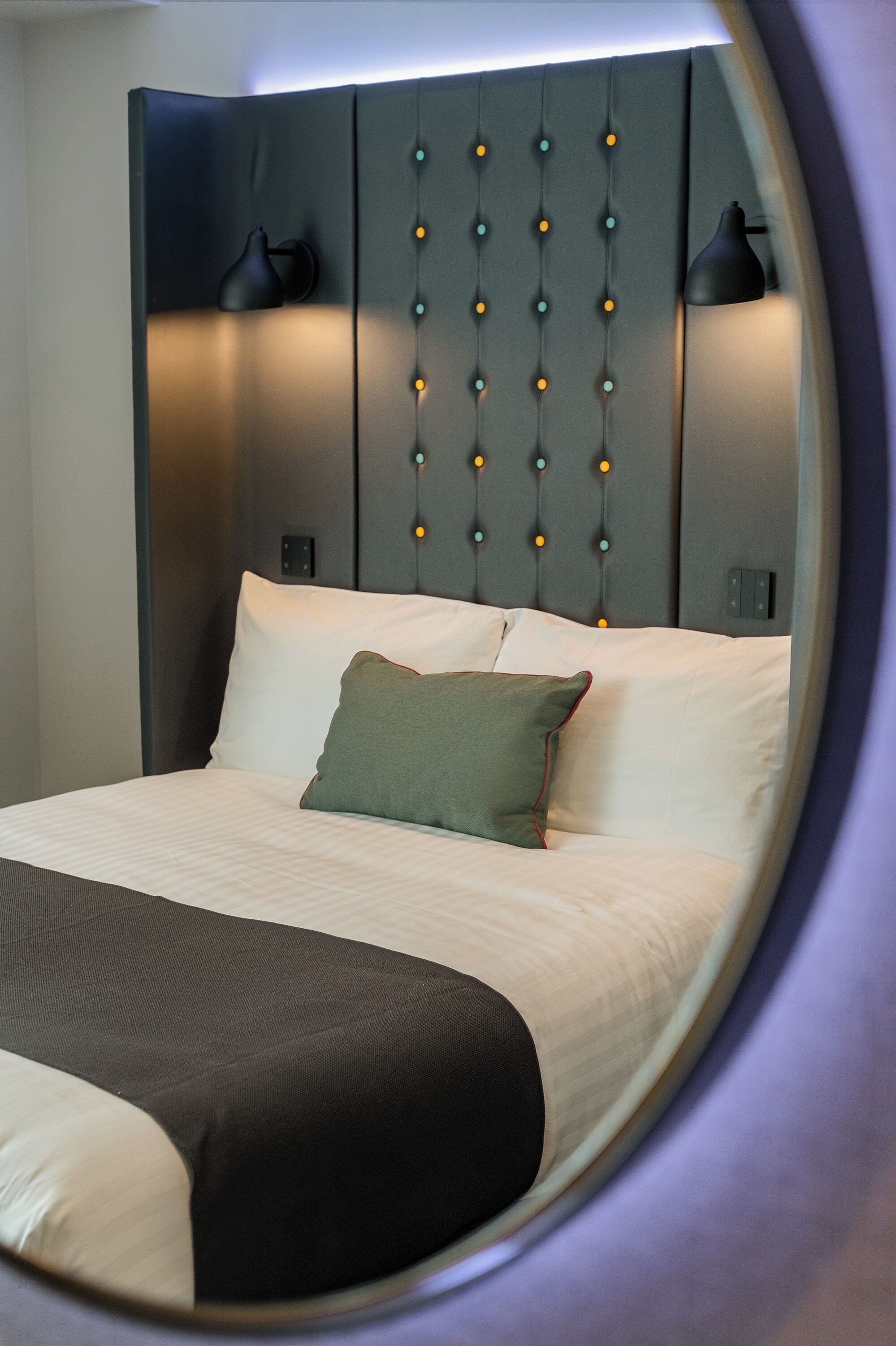 A hotel room with a bed, a headboard with lights, and a view through a circular window.