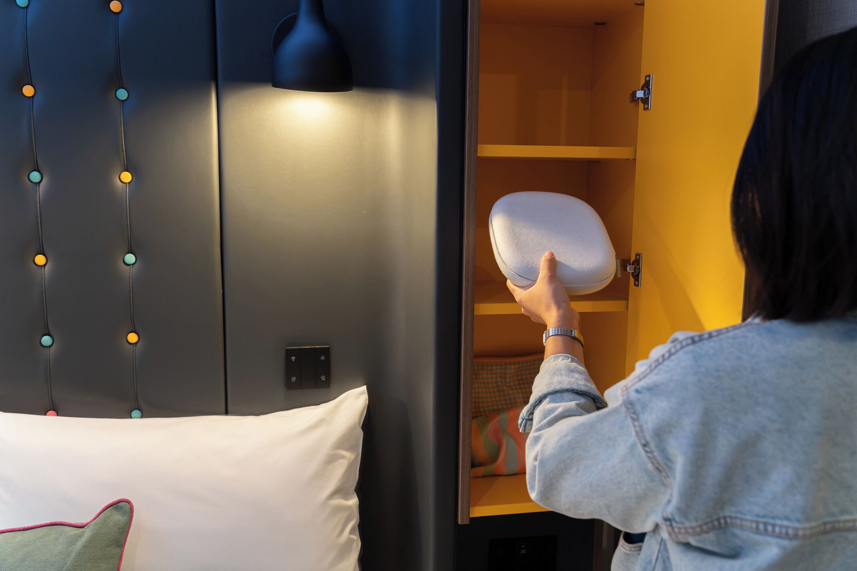 A person placing a pillow into a bed with a headboard that has a light fixture and a wall-mounted control panel.