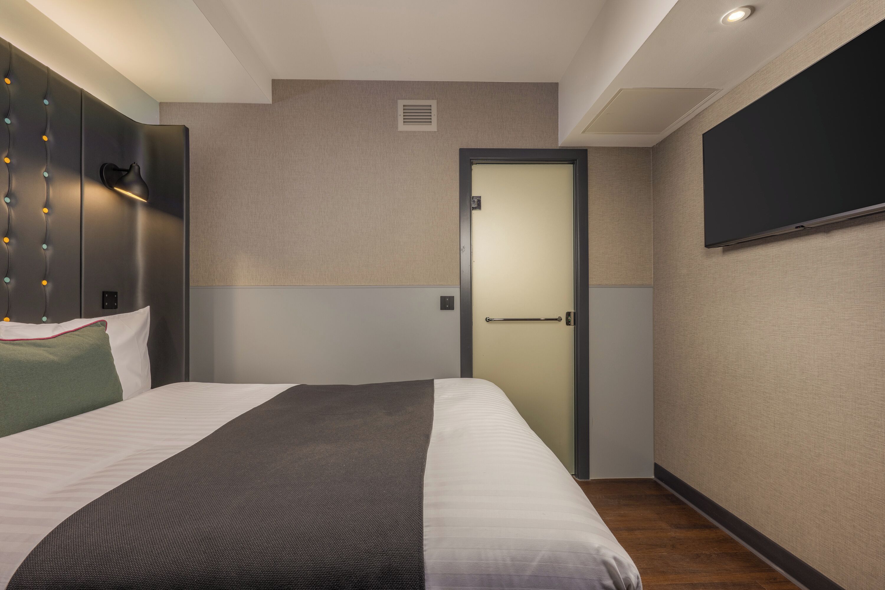 A hotel room with a bed, a wall-mounted TV, a door, and a light fixture.
